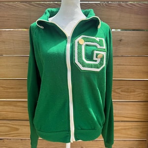 May include: Green zip-up jacket with a white and gold trim. The jacket features a large white and green letter "G" patch on the chest, with a gold basketball detail. The jacket is displayed on a mannequin.