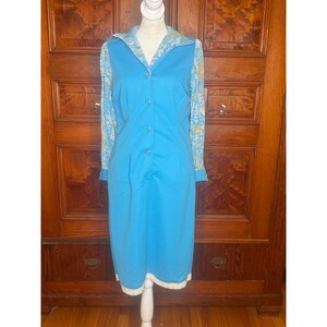 May include: A knee-length dress in bright blue with a solid blue front panel and long sleeves featuring a light blue and white floral pattern. The dress has a pointed collar, a vertical row of buttons down the center, and a white lace trim at the hem.