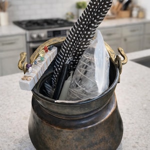 May include: A bronze cauldron with gold handles, filled with a black and white spotted feather, a white feather, a small wooden block with colorful crystals, and a clear plastic bag. The cauldron sits on a light gray countertop.