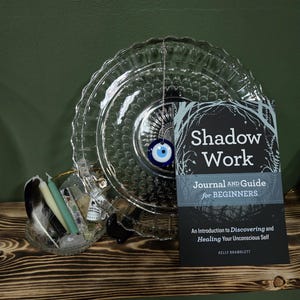 May include: A clear glass plate with a decorative edge holds a blue and white evil eye charm. A book titled "Shadow Work Journal and Guide for Beginners" is displayed next to the plate. The image also includes candles and feathers.