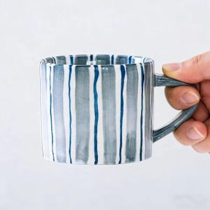 Könnte beinhalten: Eine weiße Keramik-Tasse mit schwarzem Henkel und dem Text "But First, Coffee" in fetter, schwarzer Schrift. Die Tasse steht vor einem neutralen Hintergrund, der ihr schlichtes Design hervorhebt.