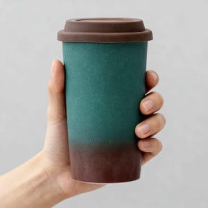 May include: A teal and brown travel mug with a brown lid is held in a hand. The mug has a textured surface and a gradient colour scheme, transitioning from teal to brown. The lid is made of a similar brown material.
