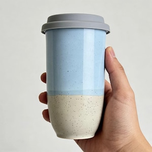 Pode incluir: Uma caneca de viagem de cerâmica de duas cores com uma tampa de silicone cinza. A metade superior é azul claro com detalhes salpicados, e a metade inferior é branco sujo com salpicos semelhantes. A caneca é segurada por uma mão.