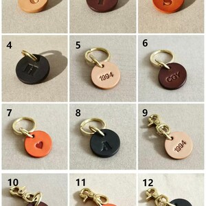 May include: A collection of twelve circular leather tags in various colors including tan, brown, orange, and black. Each tag features embossed letters, numbers, or a heart symbol and is attached to a gold-toned metal split ring or lobster clasp.