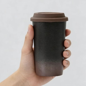 Może przedstawiać: Czarny i brązowy kubek podróżny z brązową pokrywką. Kubek jest trzymany w dłoni i ma teksturowaną powierzchnię. Dół kubka przechodzi od czarnego do brązowego.
