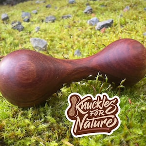 May include: A handcrafted, dark brown wooden massage tool with a unique hourglass shape, resting on a bed of green moss. The tool is smooth and polished. A sticker with the text "Knuckles for Nature" is also visible.