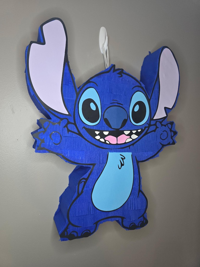 May include: A blue pi&ntilde;ata in the shape of the character Stitch, with large ears, a light blue belly, and a wide smile. The pi&ntilde;ata is hanging against a gray wall, ready for a party.