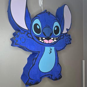 May include: A blue pi&ntilde;ata in the shape of the character Stitch, with large ears, a light blue belly, and a smiling face. The pi&ntilde;ata is hanging on a wall with a white hook.