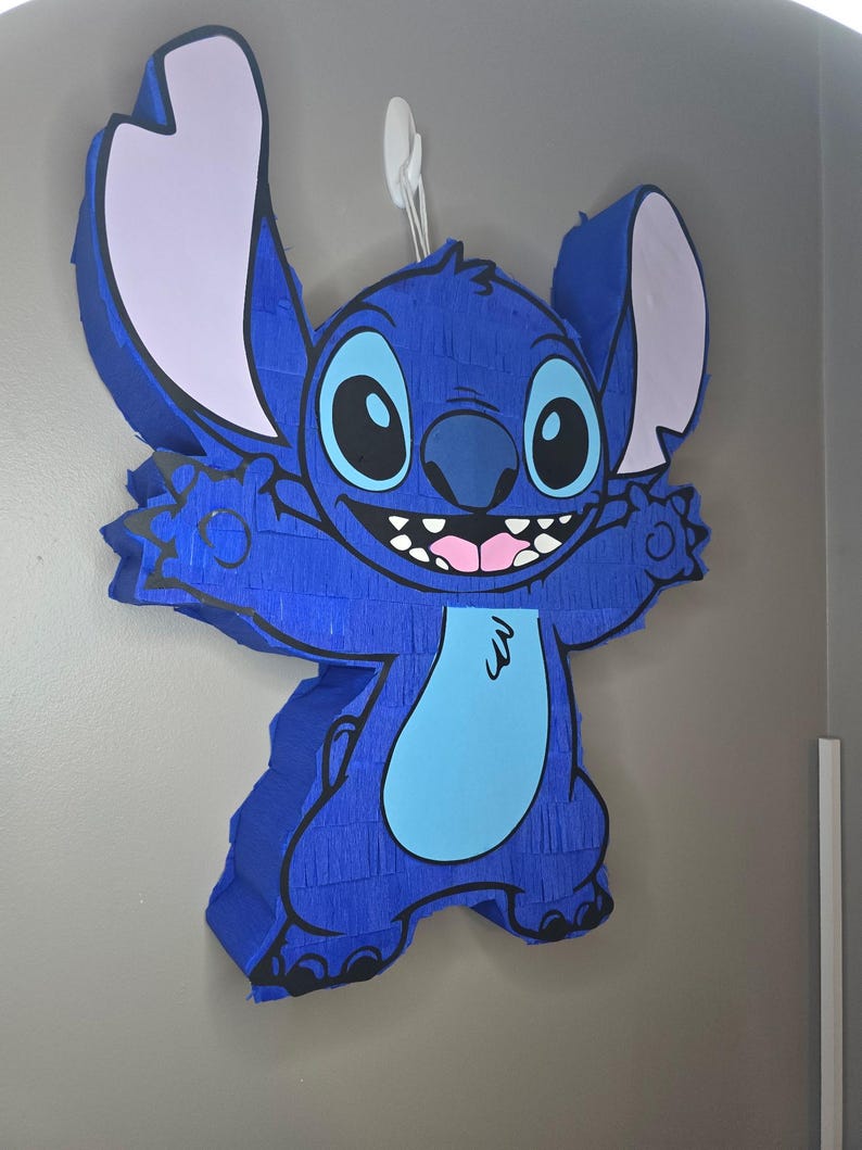 May include: A blue pi&ntilde;ata in the shape of the character Stitch, with large ears, a light blue belly, and a smiling face. The pi&ntilde;ata is hanging on a wall with a white hook.