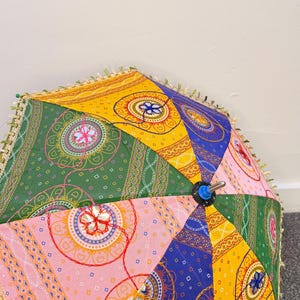 May include: A multi-colored parasol featuring a patchwork design of yellow, green, pink, and blue fabric panels. Each panel displays intricate circular mandala patterns, geometric shapes, and chain-link motifs, finished with a decorative fringe trim.
