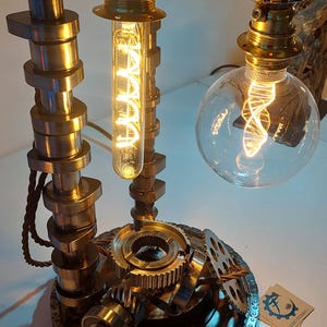 Puede incluir: Una lámpara de mesa construida con piezas de automóvil recicladas, incluyendo una base de engranajes de metal, árboles de levas y una cadena. Cuenta con dos bombillas de filamento expuesto, una tubular y otra esférica, con cableado marrón.