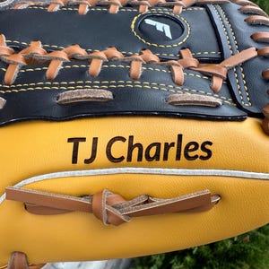 May include: Close-up of a baseball glove with a black top and yellow palm. The glove has brown leather lacing and stitching. The text "TJ Charles" is printed on the palm. A white logo is visible on the black top.