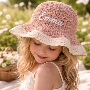 May include: A dusty rose crochet bucket hat featuring a wide, cream-colored scalloped lace trim along the brim. The front of the hat is embroidered with the name Emma in white cursive script, creating a textured and detailed appearance.