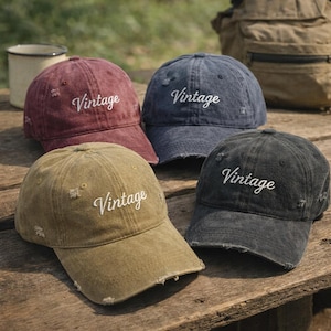 Puede incluir: Cuatro gorras de béisbol desgastadas en burdeos, azul marino, verde oliva y gris carbón. Cada gorra tiene la palabra "Vintage" bordada en blanco. Las gorras están sobre una superficie de madera.