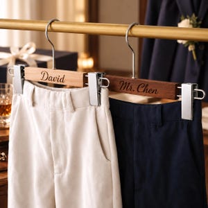 May include: Two wooden pants hangers with silver metal clips hanging from a gold rod. The light wood hanger is engraved with the name David, holding white trousers. The dark wood hanger is engraved with Mr. Chen, holding navy blue trousers.
