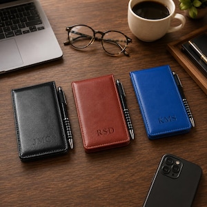 May include: Three rectangular leather-like pocket notebooks in black, brown, and blue, each featuring a debossed monogram and a black pen held in a side loop. The black notebook is marked JMC, the brown one RSD, and the blue one KMS.