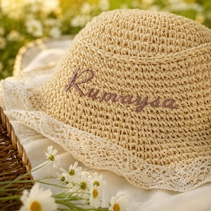 May include: A beige woven straw sun hat featuring a rounded crown and a wide brim edged with delicate white lace trim. The name Rumaysa is embroidered in brown cursive script across the front of the hat, which has a textured, open-weave construction.