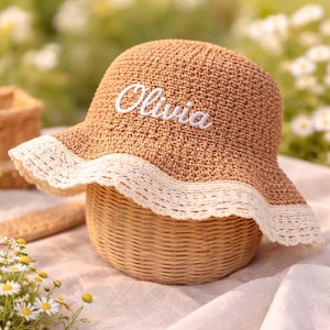 May include: A tan crocheted sun hat featuring a wide brim with a white scalloped lace trim. The front of the hat displays the name Olivia embroidered in white cursive script. The hat has a rounded crown and a textured, woven appearance.