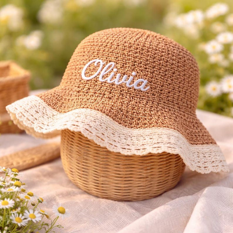 May include: A tan crocheted sun hat featuring a wide brim with a white scalloped lace trim. The front of the hat displays the name Olivia embroidered in white cursive script. The hat has a rounded crown and a textured, woven appearance.
