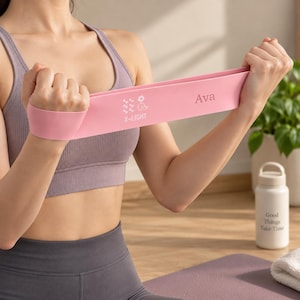 Puede incluir: Una banda de resistencia de color rosa claro hecha de material liso, con el texto impreso 'Ava' y 'X-LIGHT' junto a un pequeño icono de una flor y una mancuerna. La banda se sostiene horizontalmente, mostrando su estructura plana y flexible.