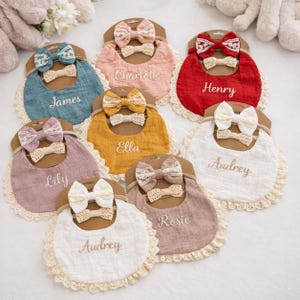 May include: A collection of eight fabric baby bibs and matching hair bows on cardboard backing. Each bib features a scalloped lace trim, a personalized embroidered name, and a coordinating bow set in colors including blue, pink, red, yellow, and white.