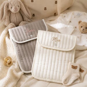 May include: A selection of baby essentials, featuring a grey and a cream quilted changing bag, each embroidered with a name. Also includes a plush bunny, a dummy, a teddy bear, a babygrow, and socks.
