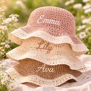 May include: A stack of three crocheted bucket hats in shades of dusty pink, cream, and tan. Each hat features a scalloped brim and a name embroidered in cursive script on the front: Emma in white, Lily in brown, and Ava in white thread.