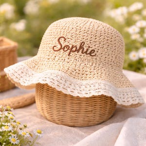 May include: A beige crocheted bucket hat featuring a wide, scalloped white lace trim along the brim. The name Sophie is embroidered in brown cursive script on the front of the hat, which has a textured, woven appearance throughout the crown.