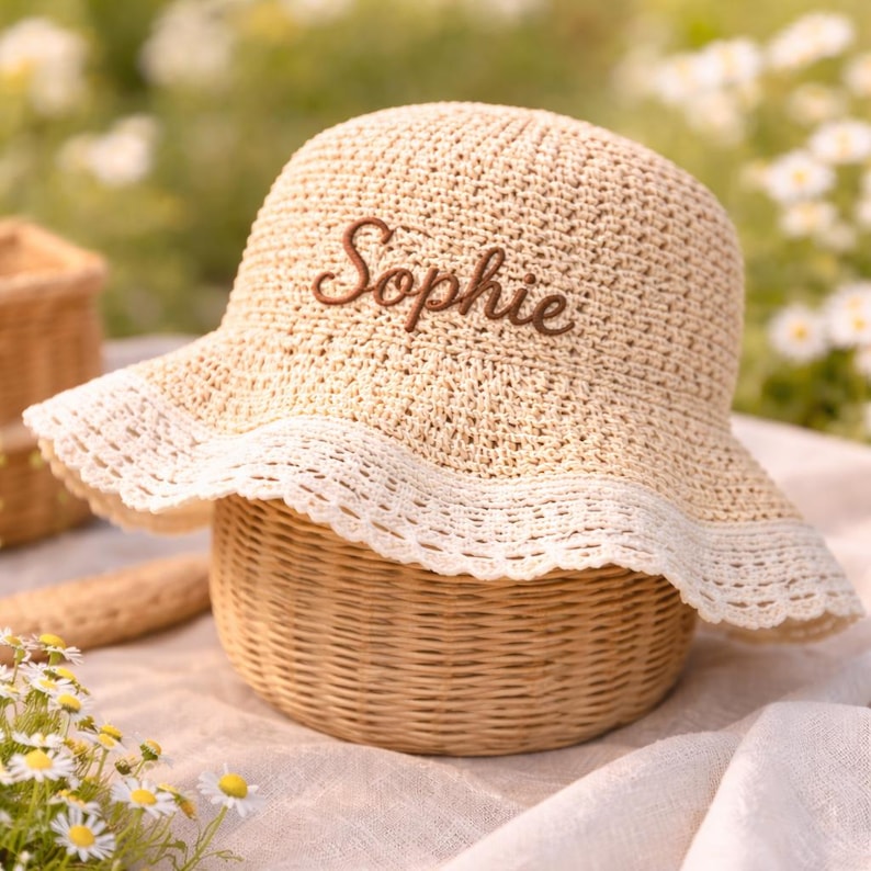 May include: A beige crocheted bucket hat featuring a wide, scalloped white lace trim along the brim. The name Sophie is embroidered in brown cursive script on the front of the hat, which has a textured, woven appearance throughout the crown.
