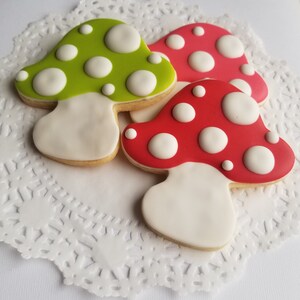 May include: Three decorated sugar cookies shaped like mushrooms. One cookie is green with white dots, one is red with white dots, and one is red with white dots. The cookies are on a white doily.