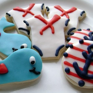 May include: A set of decorated sugar cookies featuring nautical themes. The cookies are shaped like whales, anchors, and sailor shirts. The cookies are decorated with red, white, and blue icing.