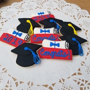 May include: A collection of graduation-themed sugar cookies decorated with red, white, and blue icing. The cookies feature graduation caps, diplomas, and the text "Congrats!" and "2019".