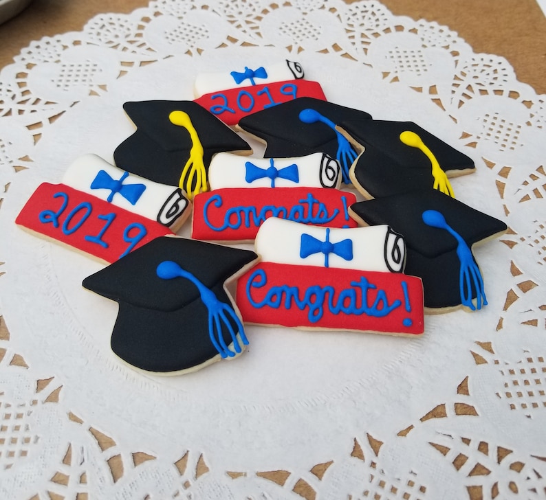 May include: A collection of graduation-themed sugar cookies decorated with red, white, and blue icing. The cookies feature graduation caps, diplomas, and the text "Congrats!" and "2019".