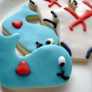 May include: Two blue whale cookies decorated with white icing and red hearts. The cookies are on a white plate.