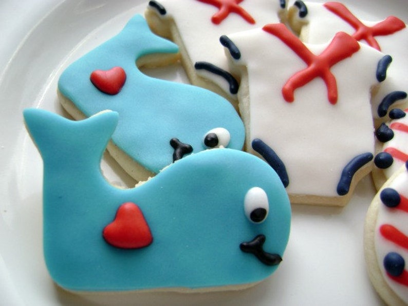 May include: Two blue whale cookies decorated with white icing and red hearts. The cookies are on a white plate.
