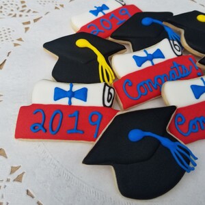 May include: Assortment of graduation themed sugar cookies decorated with blue, red, white, and yellow icing. The cookies feature a graduation cap, a diploma, and the year 2019. The cookies are decorated with the words "Congrats!" and "2019".