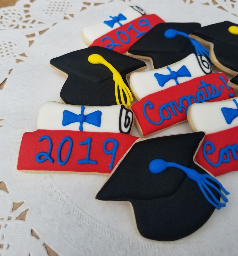 May include: Assortment of graduation themed sugar cookies decorated with blue, red, white, and yellow icing. The cookies feature a graduation cap, a diploma, and the year 2019. The cookies are decorated with the words "Congrats!" and "2019".