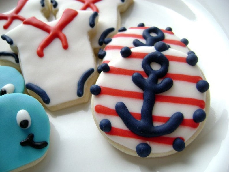 May include: A set of decorated sugar cookies with nautical themes. The cookies are decorated with red and blue icing and feature an anchor, a sailor's shirt, and a blue whale.