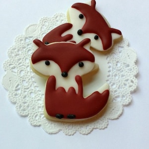 May include: Three fox-shaped sugar cookies decorated with red and white icing and black eyes. The cookies are on a white doily.