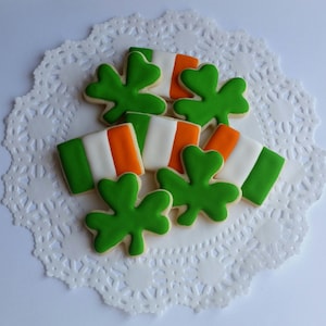 May include: A collection of St. Patrick's Day cookies. The cookies are decorated with green shamrocks and Irish flags in green, white, and orange. The cookies are arranged on a white, lacy paper doily.
