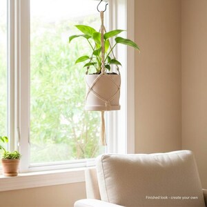 May include: A beige ceramic plant pot held by a cream-coloured macrame hanger with a diamond pattern and a bottom tassel. The hanger is suspended from a hook, containing a green leafy plant. The text "Finished look - create your own" is visible.
