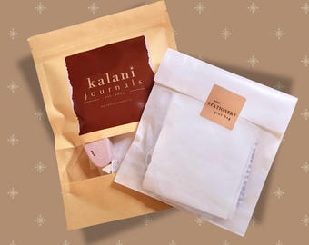 May include: Two packages of stationery products. One is a kraft paper bag with a brown label that reads "kalani journals est. 2023". The other is a white paper bag with a small sticker that reads "mini stationery grab bag".