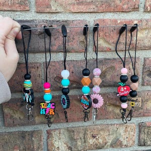 May include: A collection of beaded lanyards with various charms, including a teacher-themed lanyard with the text "Teacher" and an apple. Other lanyards feature floral, geometric, and patterned beads in vibrant colors. Each lanyard has a black cord and a metal clasp.