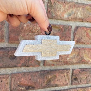 May include: A white and tan Chevrolet logo keyring. The logo is a stylized bow tie shape with a textured tan centre and a white border. The keyring is held up against a brick wall.