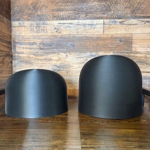 May include: Two black, dome-shaped objects of different sizes sit on a wooden surface. The objects have a smooth, matte finish and a rounded top. The background is a wooden wall.