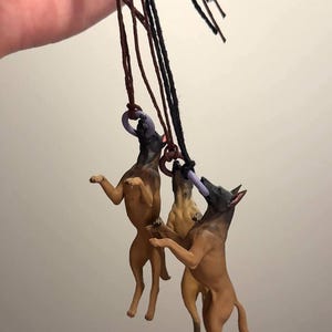 May include: Three miniature dog figurines in a playful pose, each with a brown and black coat, are suspended by black and brown cords. They are reaching for purple rings. The dogs are in a dynamic, mid-air stance.