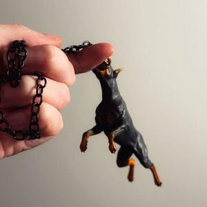 May include: A black and tan Doberman dog figurine pendant attached to a black metal link chain. The dog is depicted in a dynamic jumping pose with its front paws raised and its head tilted upward, showcasing detailed muscular features.