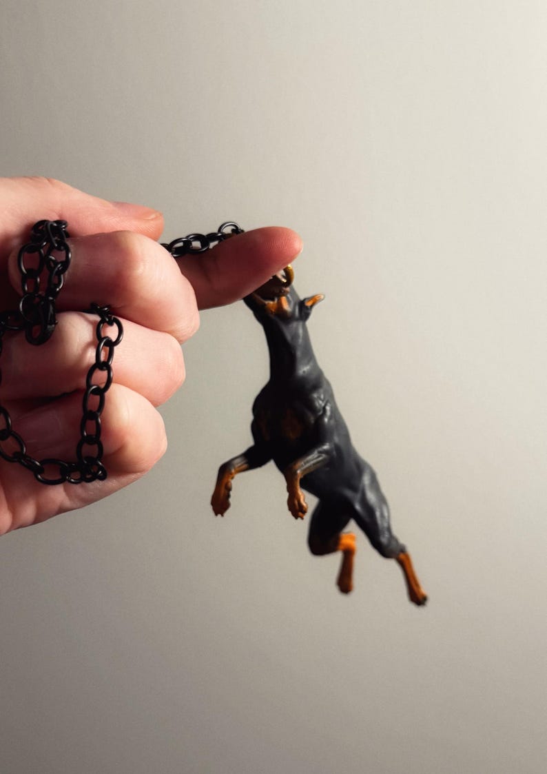 May include: A black and tan Doberman dog figurine pendant attached to a black metal link chain. The dog is depicted in a dynamic jumping pose with its front paws raised and its head tilted upward, showcasing detailed muscular features.