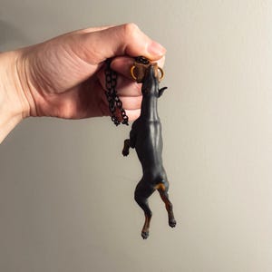 May include: A black Doberman Pinscher pendant with a gold-colored collar and a black chain. The dog is standing on its hind legs, reaching up towards a small brown object. The pendant is held up by a hand.