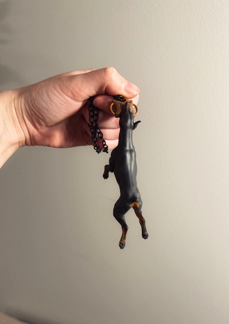 May include: A black Doberman Pinscher pendant with a gold-colored collar and a black chain. The dog is standing on its hind legs, reaching up towards a small brown object. The pendant is held up by a hand.